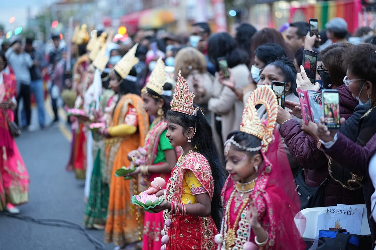Young participants in colorful ceremonial dress stand along the street as families watch the Diwali celebration.